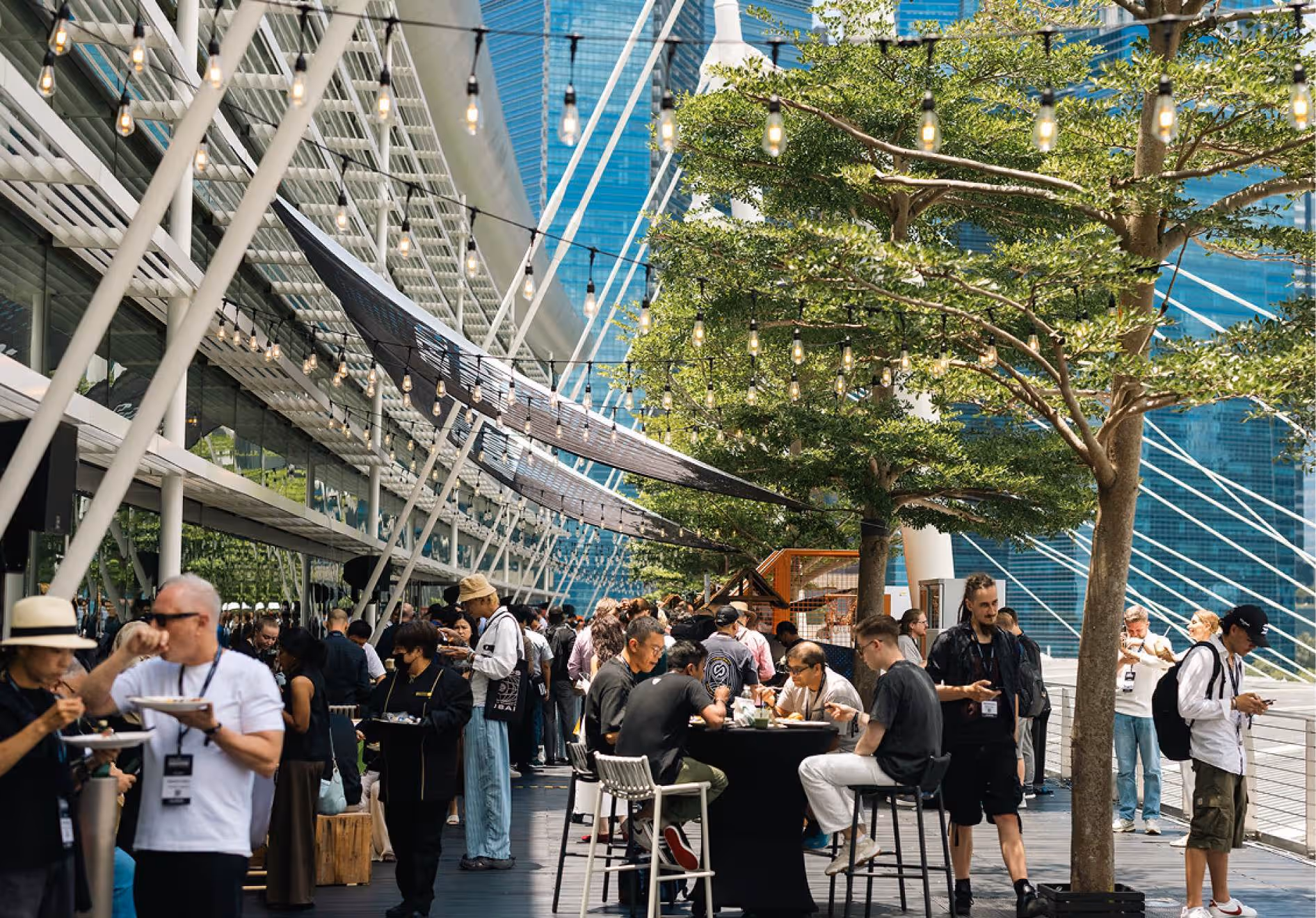 Crowd of people eating and standing at outdoor event under string lights with city buildings and trees in background.